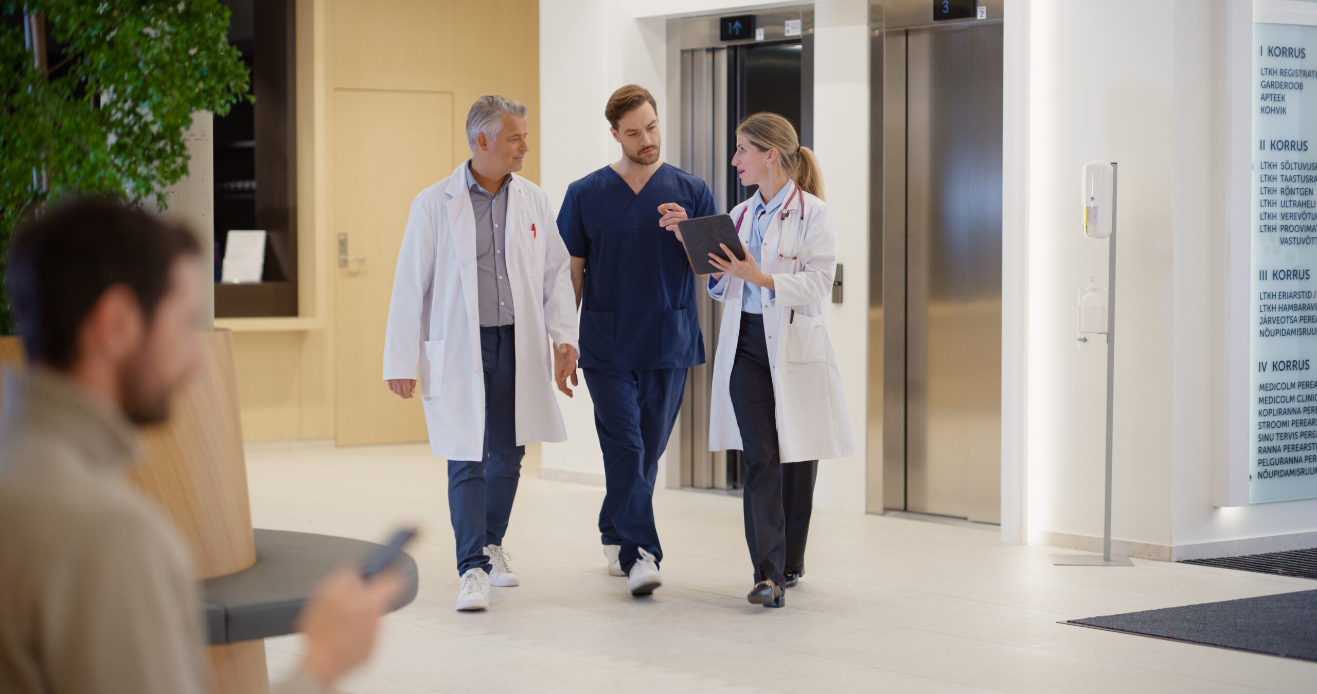 Medical Professionals Having a Discussion while Walking Through a Hospital Lobby. Middle Aged Doctor, Male Nurse in Scrubs and Female Physician with Tablet Discussing Healthcare